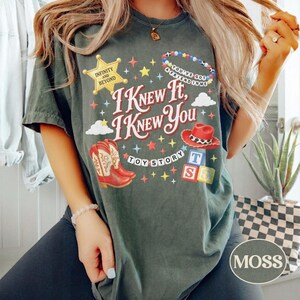 以下が含まれることがあります： I KNEW IT, I KNEW YOU, TOY STORY, INFINITY AND BEYOND、YOU'VE GOT A FRIEND IN MEのテキストがプリントされたダークグリーンのTシャツ。デザインには、黄色い保安官の星、ビーズのネックレス、赤いカウボーイハット、ブーツ、積み木が含まれています。