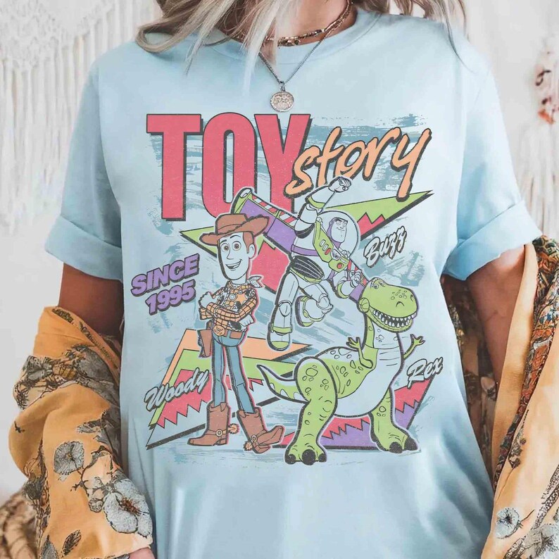 May include: Light blue t-shirt featuring a graphic design from the movie "Toy Story." The design includes Woody, Buzz Lightyear, and Rex, with the text "TOY STORY" and "SINCE 1995."