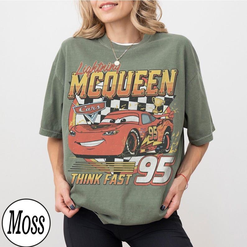 May include: Olive green t-shirt featuring a graphic of Lightning McQueen from the Cars movie. The design includes the text "Lightning McQueen", "Cars", "Think Fast", and the number "95". The shirt has a vintage, distressed look.