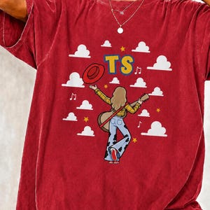 May include: A red short-sleeved t-shirt featuring a graphic of a person with long hair wearing a yellow plaid shirt and blue cow-print bell-bottom trousers, holding a guitar and a red hat. The design includes white clouds, yellow stars, and the text TS.