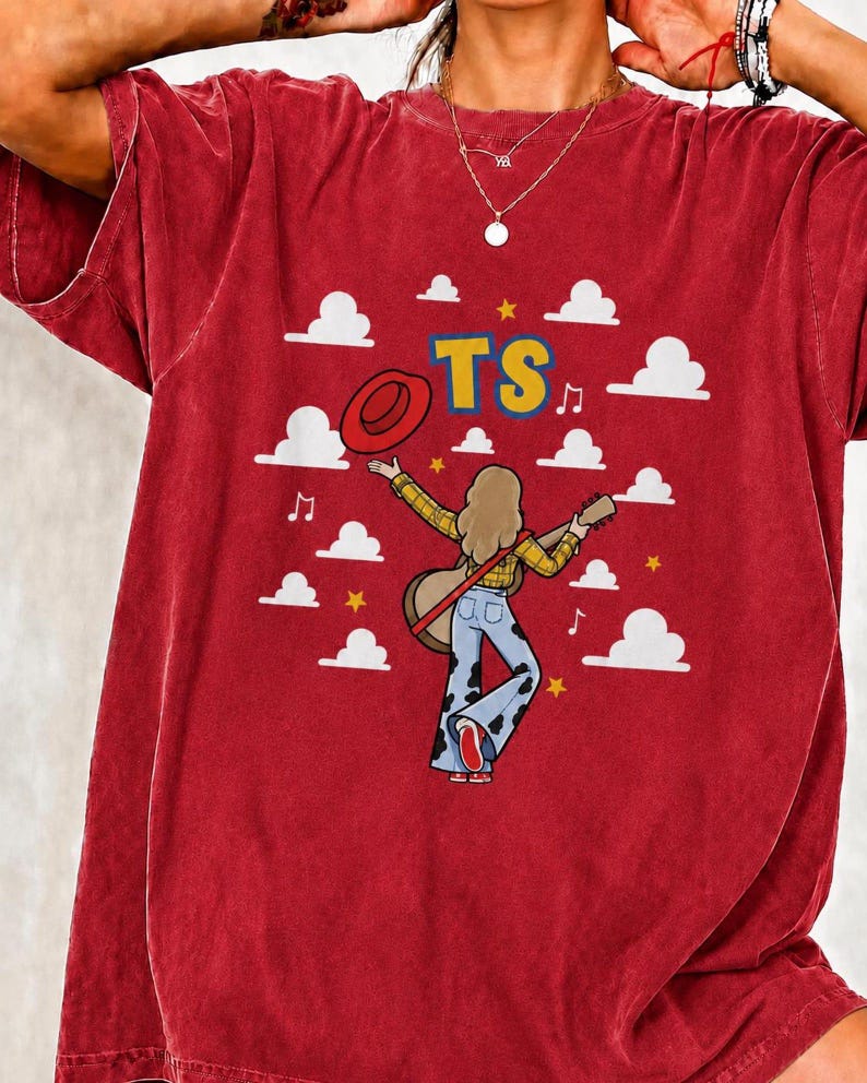 May include: A red short-sleeved t-shirt featuring a graphic of a person with long hair wearing a yellow plaid shirt and blue cow-print bell-bottom trousers, holding a guitar and a red hat. The design includes white clouds, yellow stars, and the text TS.