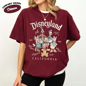 K&ouml;nnte beinhalten: Karmesinrotes T-Shirt mit wei&szlig;em Kragen und Disneyland-Grafik. Die Grafik zeigt das Disneyland-Logo, den Text "California" und Zeichentrickfiguren wie Mickey Mouse und Donald Duck. Das Shirt ist von der Marke Comfort Colors.