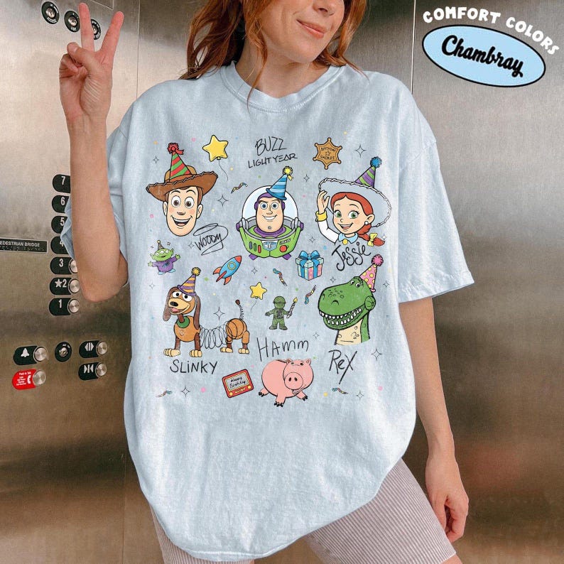 May include: A light blue t-shirt featuring a graphic print of Toy Story characters including Woody, Buzz Lightyear, Jessie, Slinky Dog, Hamm, and Rex. The characters are wearing party hats, and the text includes their names and the phrase Comfort Colors Chambray.