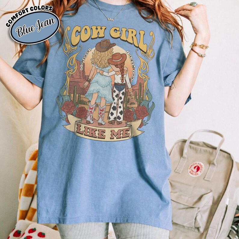 Puede incluir: Una camiseta de color azul vaquero con un estampado gr&aacute;fico de dos figuras con sombreros de vaquero en un paisaje des&eacute;rtico con cactus y rosas. El texto COW GIRL est&aacute; impreso sobre las figuras y LIKE ME aparece en una cinta debajo.