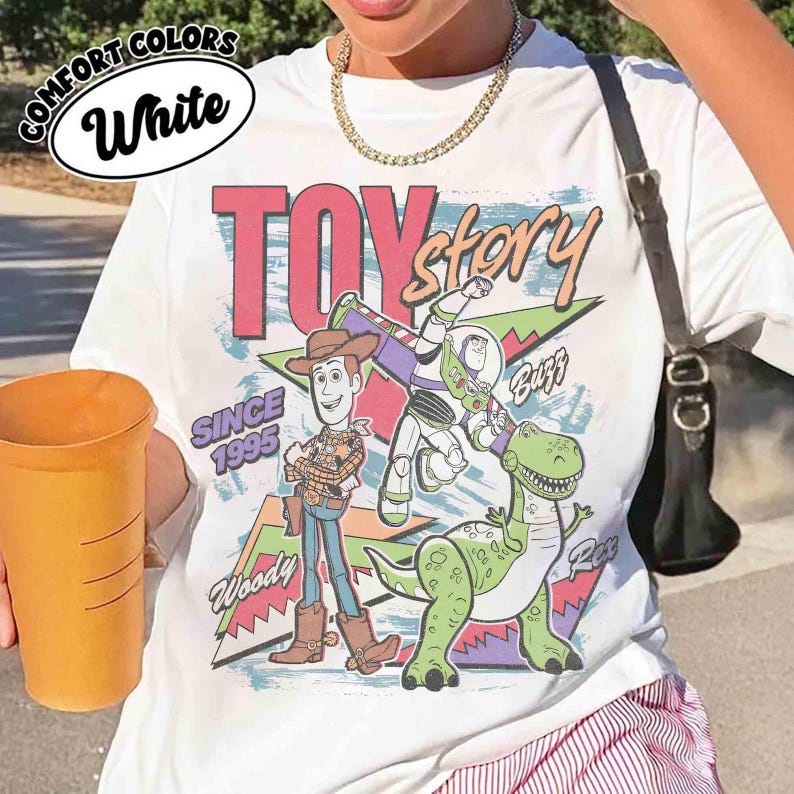 May include: White t-shirt featuring a colorful graphic of Toy Story characters Woody, Buzz Lightyear, and Rex. The design includes the text "TOY STORY" and "SINCE 1995". The shirt has the words "COMFORT COLORS" and "White" in a circle.