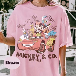 Może przedstawiać: Jasnor&oacute;żowa koszulka z grafiką postaci Disneya, w tym Myszki Miki, Myszki Minnie, Kaczora Donalda i Goofy'ego. Projekt zawiera napis "MICKEY & CO. EST 1928" i "Lightning McQueen". Na koszulce wydrukowano r&oacute;wnież słowo "Blossom".