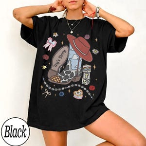 Puede incluir: Una camiseta negra de manga corta con un estampado gr&aacute;fico de una bota de vaquero con patr&oacute;n de vaca, un sombrero rojo, un fondo de cielo azul y bloques apilados con las letras T, S y S. El texto Toy's Story est&aacute; impreso en la bota.