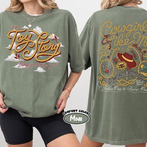 May include: A moss green t-shirt featuring a front graphic with the text "it's a Toy Story Baby just say yes" in rope-style lettering over clouds. The back graphic displays "Cowgirl like me" and "Takes One to Know One" with a hat and lasso.