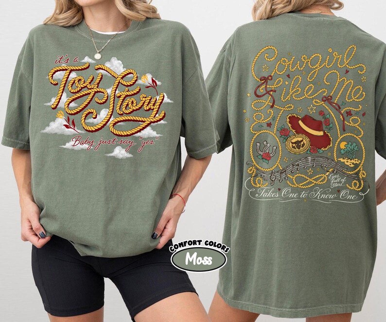 May include: A moss green t-shirt featuring a front graphic with the text "it's a Toy Story Baby just say yes" in rope-style lettering over clouds. The back graphic displays "Cowgirl like me" and "Takes One to Know One" with a hat and lasso.