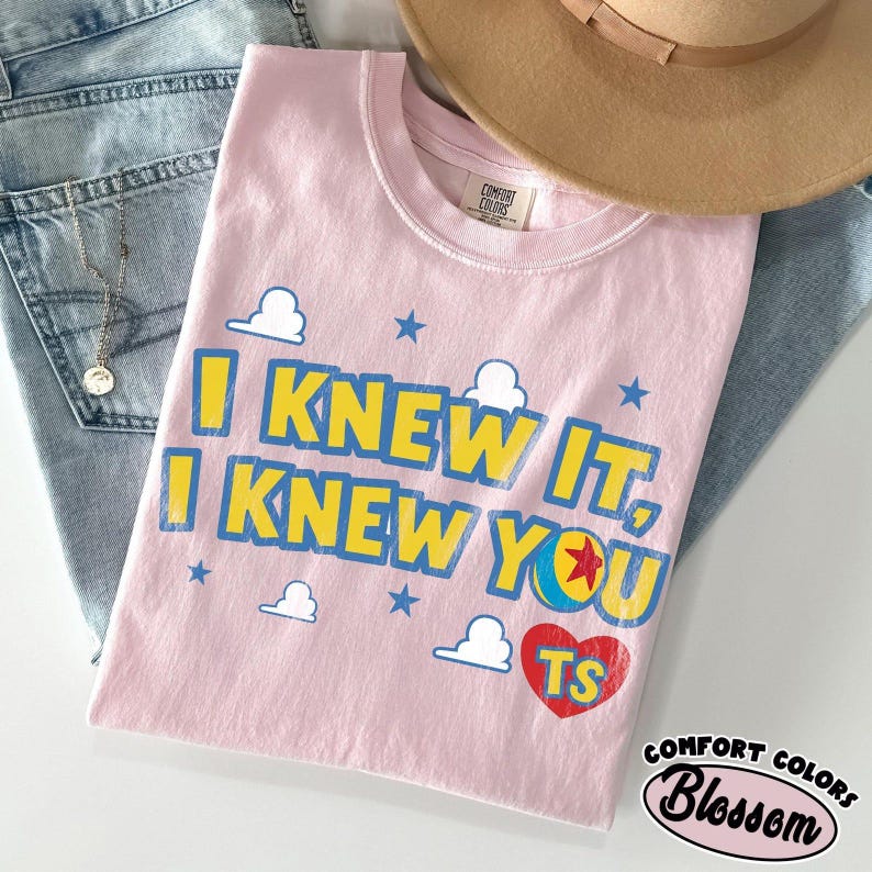 Może przedstawiać: Jasnor&oacute;żowy T-shirt z kr&oacute;tkim rękawem z ż&oacute;łto-niebieskim napisem I KNEW IT, I KNEW YOU oraz czerwonym sercem z literami TS. Wz&oacute;r zawiera małe niebieskie gwiazdki i białe chmurki. Koszulka posiada metkę z napisem Comfort Colors.