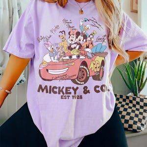 Może przedstawiać: Jasnofioletowa koszulka z grafiką postaci Disneya, w tym Myszki Miki, Myszki Minnie, Kaczora Donalda i Goofy'ego, w czerwonym samochodzie wyścigowym. Tekst "Mickey & Co. EST 1928" jest wydrukowany poniżej.