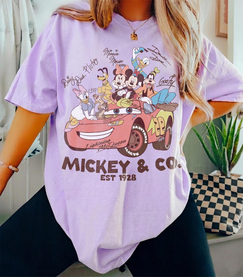 Może przedstawiać: Jasnofioletowa koszulka z grafiką postaci Disneya, w tym Myszki Miki, Myszki Minnie, Kaczora Donalda i Goofy'ego, w czerwonym samochodzie wyścigowym. Tekst "Mickey & Co. EST 1928" jest wydrukowany poniżej.