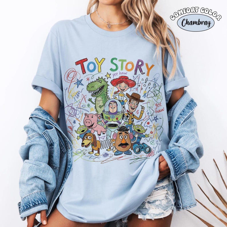 May include: A light blue short-sleeve t-shirt featuring a colorful graphic print of Toy Story characters including Woody, Buzz Lightyear, Jessie, Rex, Hamm, Slinky Dog, Mr. Potato Head, and Aliens, surrounded by multi-colored scribbles and text.