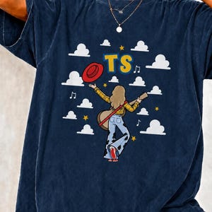 May include: A dark blue t-shirt featuring a graphic of a person with long hair wearing a yellow plaid shirt and cow-print bell-bottom trousers, holding a guitar and a red hat. The design includes white clouds, yellow stars, music notes, and the text TS.