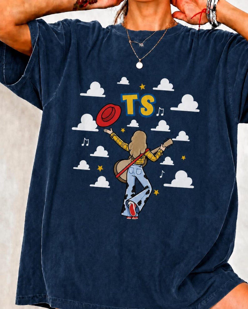 May include: A dark blue t-shirt featuring a graphic of a person with long hair wearing a yellow plaid shirt and cow-print bell-bottom trousers, holding a guitar and a red hat. The design includes white clouds, yellow stars, music notes, and the text TS.