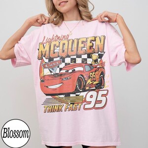May include: A light pink t-shirt featuring a graphic of Lightning McQueen from the Cars movie. The design includes the text "Lightning McQueen", "Cars", "Think Fast", and the number "95".