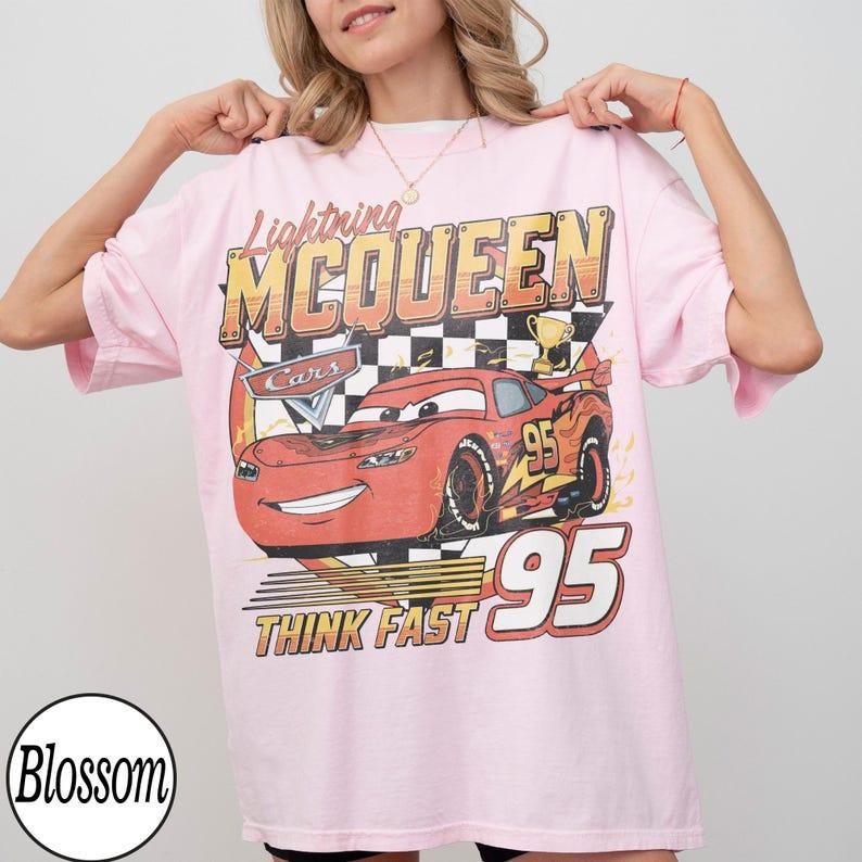 May include: A light pink t-shirt featuring a graphic of Lightning McQueen from the Cars movie. The design includes the text "Lightning McQueen", "Cars", "Think Fast", and the number "95".