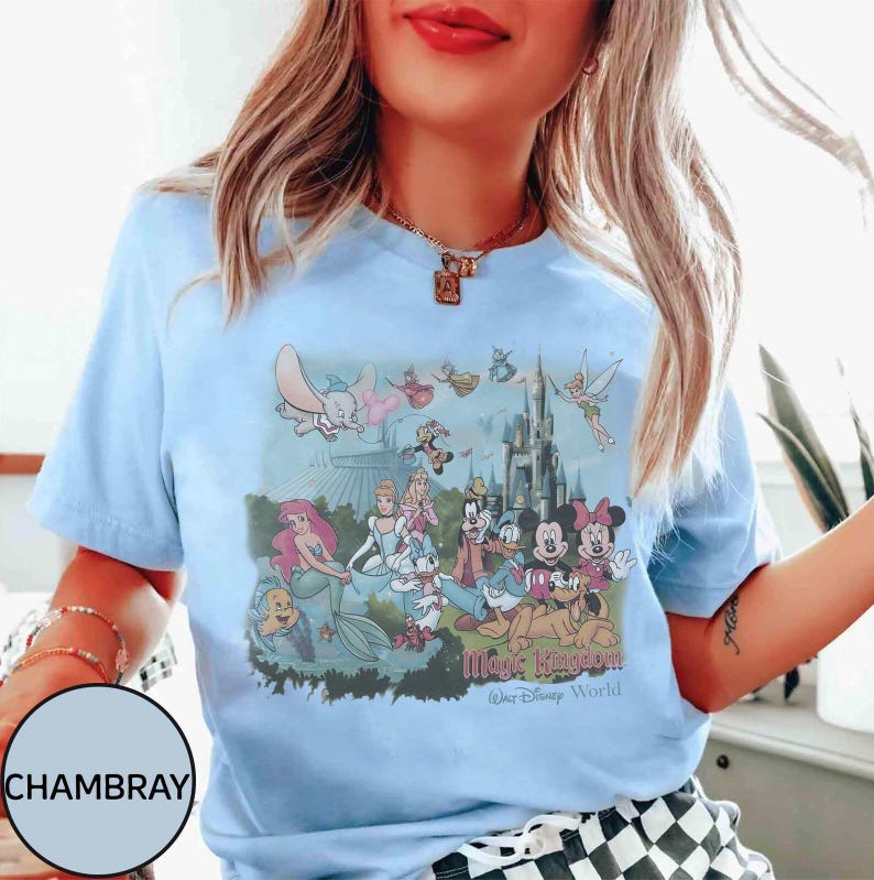 May include: Pale blue t-shirt with a vibrant graphic of Disney characters, such as Mickey and Minnie Mouse, and Ariel, set against a castle backdrop. The text reads "Magic Kingdom Walt Disney World" below the image.