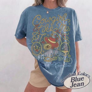 May include: A blue t-shirt featuring a graphic print with yellow rope designs, a red cowboy hat, musical notes, and the text "Cowgirl Like Me", "Eyes full of stars...", and "One to Know One". The shirt has a short-sleeved, crew-neck design.