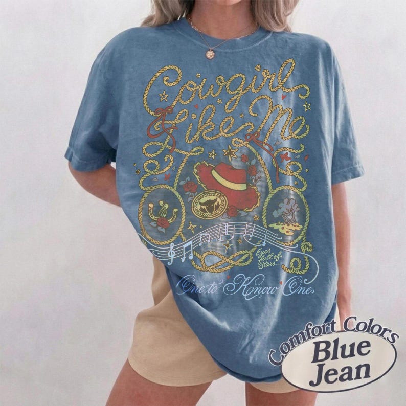 May include: A blue t-shirt featuring a graphic print with yellow rope designs, a red cowboy hat, musical notes, and the text "Cowgirl Like Me", "Eyes full of stars...", and "One to Know One". The shirt has a short-sleeved, crew-neck design.