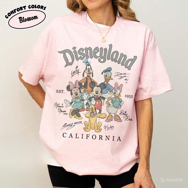K&ouml;nnte beinhalten: Ein hellrosa T-Shirt mit dem Wort "Disneyland" in einer stilisierten Schriftart sowie Zeichentrickfiguren wie Mickey Mouse, Minnie Mouse und Donald Duck. Das Shirt enth&auml;lt auch den Text "California" und "1955".