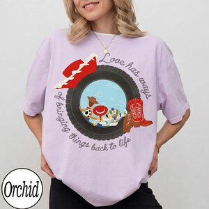 May include: A light purple t-shirt featuring a graphic of a black tire surrounding a blue sky scene with three cartoon characters. A red cowboy hat and a red cowboy boot are positioned on the tire. The text reads Love has ways of bringing things back to life.