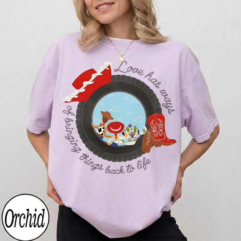 May include: A light purple t-shirt featuring a graphic of a black tire surrounding a blue sky scene with three cartoon characters. A red cowboy hat and a red cowboy boot are positioned on the tire. The text reads Love has ways of bringing things back to life.