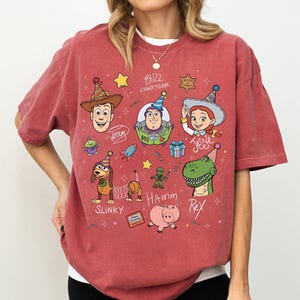 May include: A brick red t-shirt featuring a graphic print of Toy Story characters including Woody, Buzz Lightyear, Jessie, Slinky Dog, Hamm, and Rex wearing party hats. The design includes text labels for each character and small festive icons.
