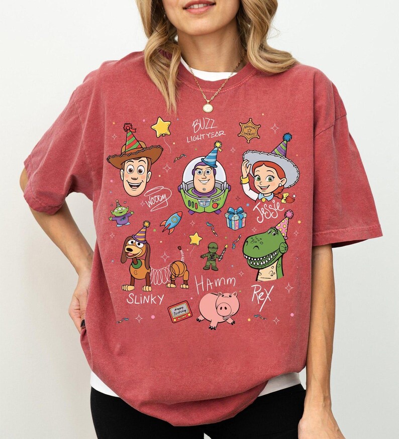 May include: A brick red t-shirt featuring a graphic print of Toy Story characters including Woody, Buzz Lightyear, Jessie, Slinky Dog, Hamm, and Rex wearing party hats. The design includes text labels for each character and small festive icons.