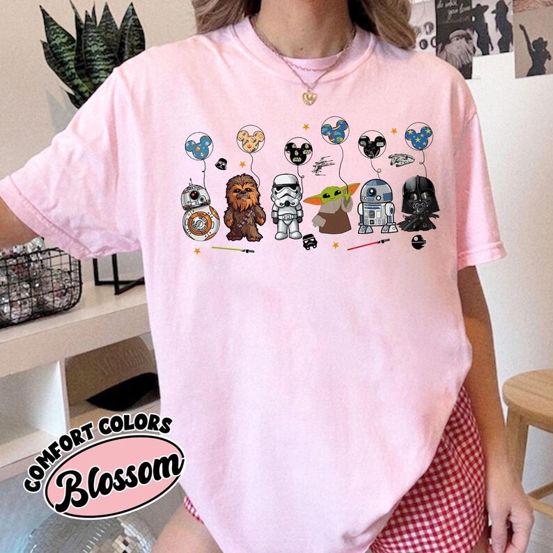 May include: A light pink Comfort Colors t-shirt featuring a graphic of Star Wars characters with Mickey Mouse-shaped balloons. The characters include BB-8, Chewbacca, a Stormtrooper, Grogu, R2-D2, and Darth Vader. The word "Blossom" is printed on the bottom left.