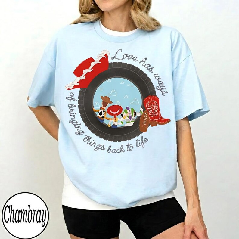 May include: A light blue t-shirt featuring a graphic of a black tire framing a sky-blue background with cartoon characters. A red cowboy hat and a red cowboy boot are positioned on the tire. The text reads, Love has ways of bringing things back to life.