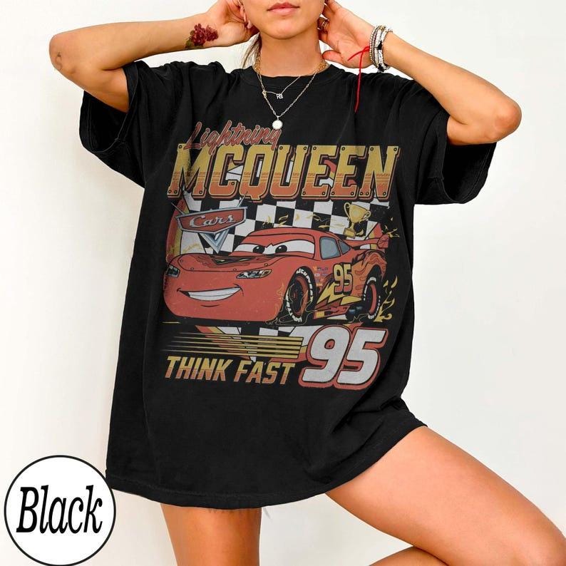 May include: Black t-shirt featuring a graphic of Lightning McQueen from the Cars movie. The design includes the text "Lightning McQueen", "Cars", "Think Fast", and the number "95". The graphic is in shades of red, yellow, and brown.