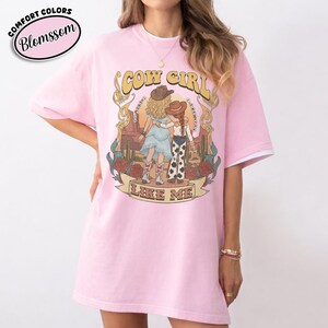 Puede incluir: Una camiseta rosa claro con un estampado gr&aacute;fico de dos figuras con sombreros de vaquero en un paisaje des&eacute;rtico con cactus y rosas. El texto "COW GIRL" aparece sobre las figuras y "LIKE ME" est&aacute; impreso en una cinta debajo de ellas.