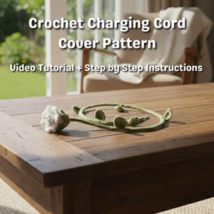 May include: A crochet charging cord cover pattern is displayed on a wooden table. The cover is green with leaf-like embellishments and a white flower-shaped end. Text on the image reads "Crochet Charging Cord Cover Pattern" and "Video Tutorial + Step by Step Instructions."