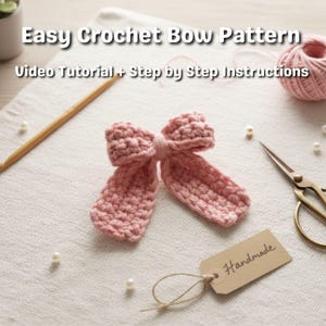 May include: A light pink crocheted bow sits on a white surface, alongside a ball of pink yarn, scissors, a wooden knitting needle, and a tag that reads "Handmade". The text "Easy Crochet Bow Pattern" is displayed above the bow. The image suggests a craft project.
