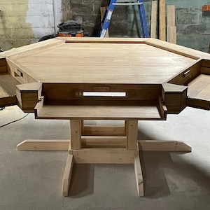Könnte beinhalten: Ein großer, unfertiger Spieltisch aus Holz mit achteckiger Spielfläche. Er verfügt über eingebaute Ablagen und einen zentralen, vertieften Bereich. Der Tisch wird von einem stabilen, hellfarbigen Holzsockel getragen, ideal für Brettspiele.