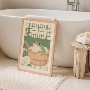 Puede incluir: Un cuadro enmarcado con un dibujo de una rana verde relajándose en una bañera de madera llena de burbujas. La obra de arte tiene un marco de madera clara y está ambientada en un baño con una bañera blanca y un pequeño taburete de madera.