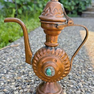 May include: A copper-colored, ornate teapot with a curved spout and a handle. The teapot features a decorative lid and a central circular design with a green stone. The teapot is standing on a small pedestal base.