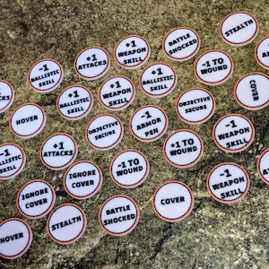 May include: A collection of white, circular game tokens with black text and a salmon-colored border. The tokens feature phrases like "+1 Attacks", "-1 Weapon Skill", and "Stealth". The tokens are scattered on a textured, green surface.