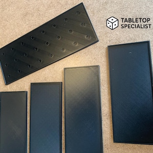 Op de afbeelding: Zwarte 3D-geprinte accessoires voor bordspellen, waaronder rechthoekige trays en een groter schuin stuk met cirkelvormige inkepingen. Het logo "Tabletop Specialist" staat rechtsboven.