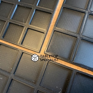 Op de afbeelding: Close-up van verschillende zwarte, vierkante roosterbakken. Elke vierkant heeft een getextureerd oppervlak. De bakken zijn gerangschikt op een lichtbruin oppervlak. De tekst "TABLETOP SPECIALIST" is zichtbaar in de rechter benedenhoek.