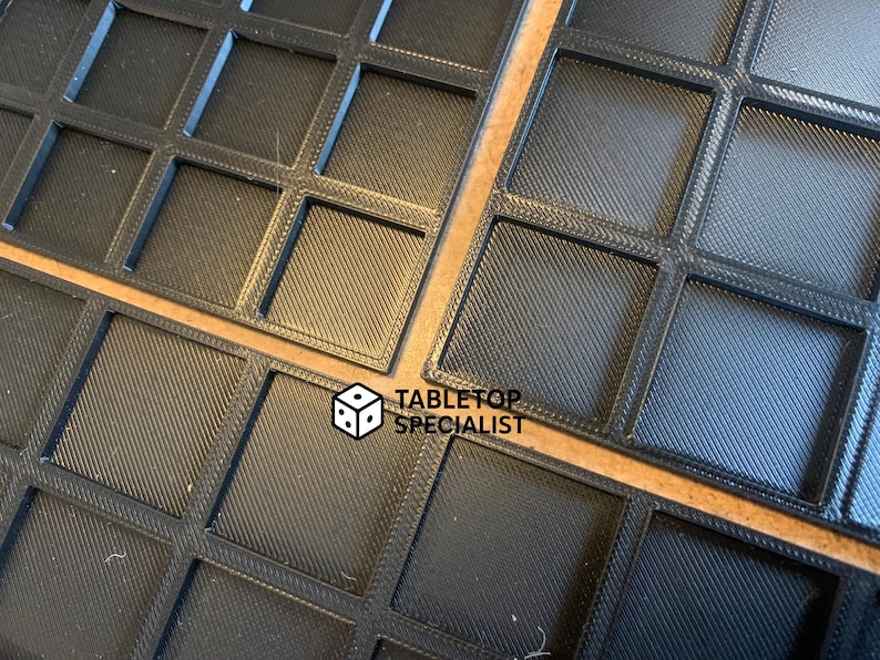 Op de afbeelding: Close-up van verschillende zwarte, vierkante roosterbakken. Elke vierkant heeft een getextureerd oppervlak. De bakken zijn gerangschikt op een lichtbruin oppervlak. De tekst "TABLETOP SPECIALIST" is zichtbaar in de rechter benedenhoek.