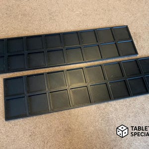 Op de afbeelding: Twee zwarte rechthoekige trays met een raster van vierkante compartimenten. De trays zijn gemaakt van een mat materiaal en zijn ontworpen voor het organiseren van kleine items. Het Tabletop Specialist-logo staat rechtsonder.