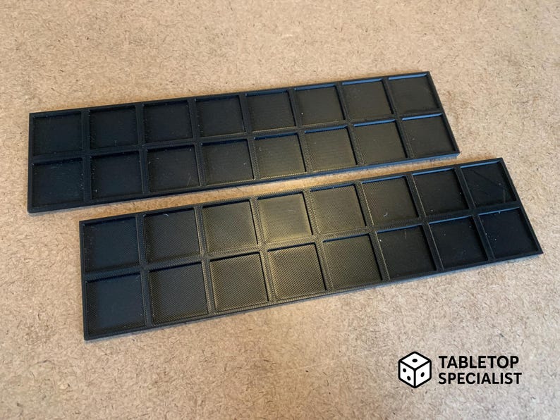 Op de afbeelding: Twee zwarte rechthoekige trays met een raster van vierkante compartimenten. De trays zijn gemaakt van een mat materiaal en zijn ontworpen voor het organiseren van kleine items. Het Tabletop Specialist-logo staat rechtsonder.