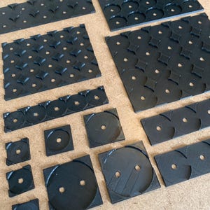 以下が含まれることがあります： 黒色の、質感のあるゲームボードコンポーネント。円形やひし形の切り抜きがある正方形と長方形のパーツが含まれています。パーツは薄茶色の表面に配置されています。Tabletop Specialistのロゴがあります。