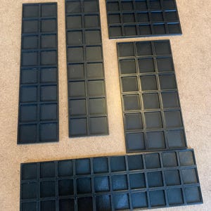 Op de afbeelding: Zwarte, modulaire tabletop-speltegels in verschillende rechthoekige en vierkante vormen. De tegels zijn gerangschikt op een lichtbruin oppervlak. Het logo van Tabletop Specialist staat rechtsonder.