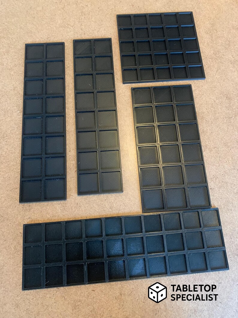 Op de afbeelding: Zwarte, modulaire tabletop-speltegels in verschillende rechthoekige en vierkante vormen. De tegels zijn gerangschikt op een lichtbruin oppervlak. Het logo van Tabletop Specialist staat rechtsonder.