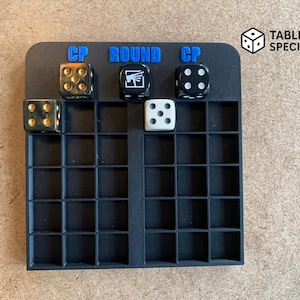 Peut inclure: Un organisateur de jeu de société noir avec plusieurs compartiments carrés. Des dés de différentes couleurs et motifs sont placés sur l'organisateur. Les mots "CP ROUND CP" sont en lettres bleues en haut. Le logo "Tabletop Specialist" est en haut à droite.