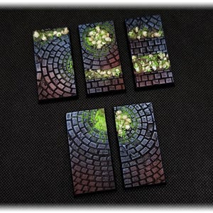 May include: A collection of rectangular, miniature terrain pieces. Each piece features a cobblestone pattern in shades of gray, brown, and red, with a circular design and green and white accents. The pieces are arranged on a black surface.