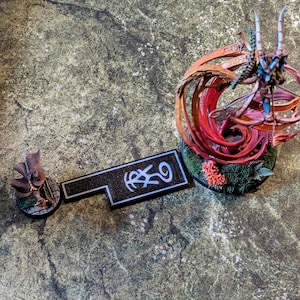 May include: Miniature fantasy figures and a black rectangular game piece with white symbols. The figures include a character with a cloak and a detailed creature with horns and a swirling, colorful design. The game piece has a white border.